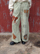Front view of the Gilded Wishes Holiday Pants sample, showing patterned fabric, bow and leaf details, pockets and distressed accents. Finished ribbon will include gold thread and crystals not shown on this sample.